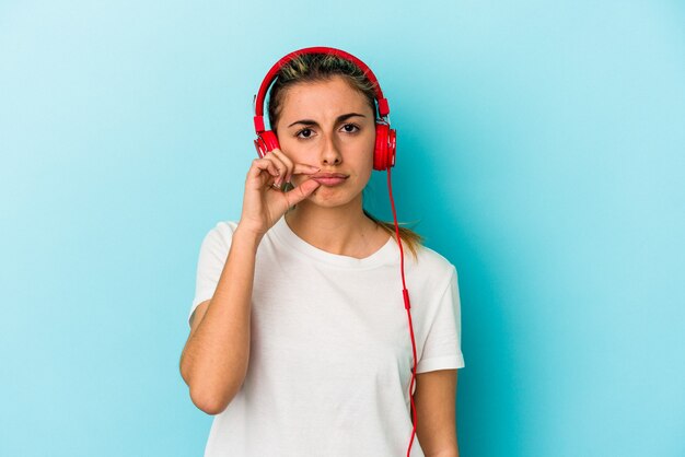Junge blonde Frau, die Musik über Kopfhörer hört, die auf blauem Hintergrund mit Fingern auf den Lippen lokalisiert wird, die ein Geheimnis halten.