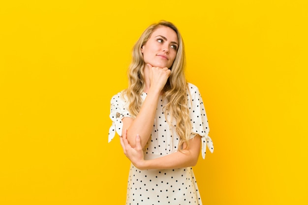 Junge blonde Frau, die nachdenklich fühlt, sich wundert oder Ideen vorstellt, Tagträumen und aufschaut, um Raum gegen gelbe Wand zu kopieren