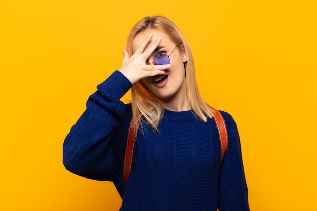 Junge blonde Frau, die schockiert, ängstlich oder verängstigt aussieht, Gesicht mit Hand bedeckt und zwischen Fingern späht