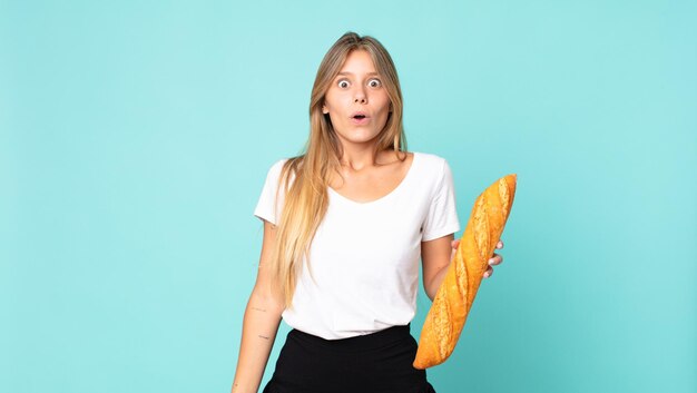 Junge blonde Frau, die sehr schockiert oder überrascht aussieht und ein Brotbaguette hält