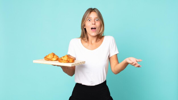 junge blonde Frau, die sich extrem schockiert und überrascht fühlt und ein Croissant-Tablett hält