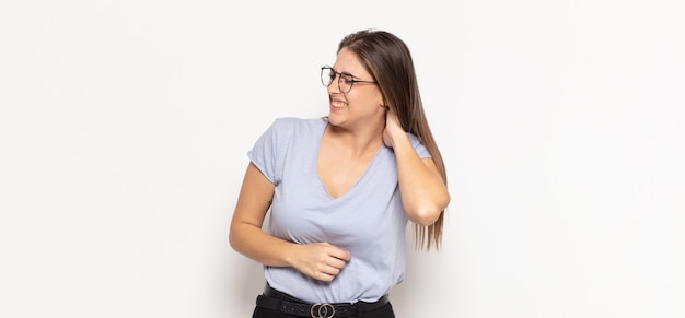 Junge blonde Frau, die sich gestresst, frustriert und müde fühlt, sich den schmerzhaften Nacken reibt, mit einem besorgten, unruhigen Blick