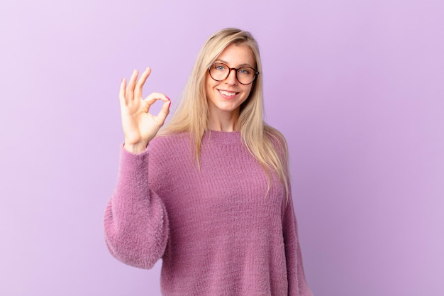 Junge blonde Frau, die sich glücklich fühlt und Zustimmung mit einer okayen Geste zeigt