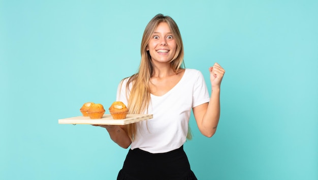 Junge blonde Frau, die sich schockiert fühlt, lacht und den Erfolg feiert und einen Muffins-Troy hält