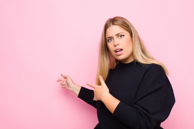 Junge blonde Frau, die sich schockiert und überrascht fühlt und auf den Kopierraum an der Seite mit erstauntem Blick mit offenem Mund gegen die flache Wand zeigt