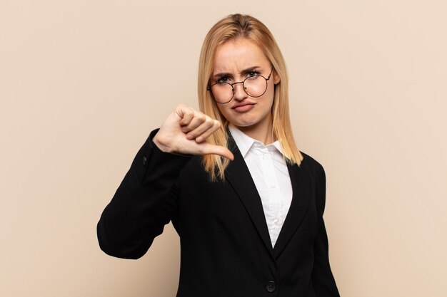 Junge blonde Frau, die sich verärgert, wütend, verärgert, enttäuscht oder unzufrieden fühlt und mit ernstem Blick Daumen nach unten zeigt