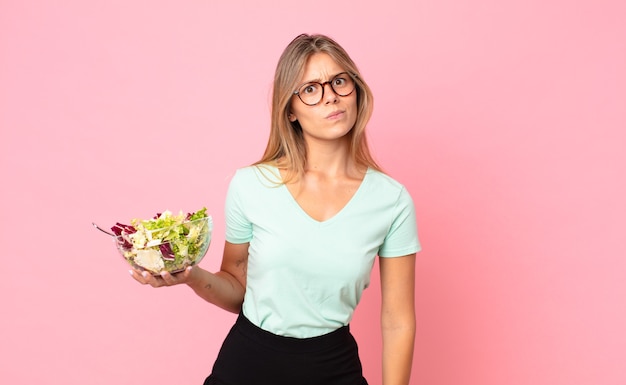 Junge blonde Frau, die traurig, verärgert oder wütend ist und zur Seite schaut und einen Salat hält