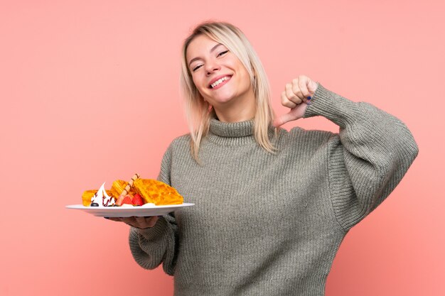 Junge blonde Frau, die Waffeln über isolierte rosa Wand stolz und selbstzufrieden hält