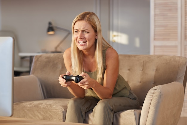 Junge blonde Frau, die zu Hause auf Sofa sitzt und Computerspiel mit Joystick spielt