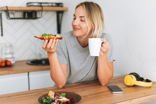 Junge blonde Frau, die zu Hause mit gesunden Sandwiches, Croissants und Tee-Morgenkaffee frühstückt