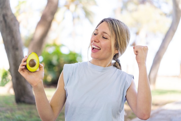 Junge blonde Frau hält draußen eine Avocado und feiert einen Sieg