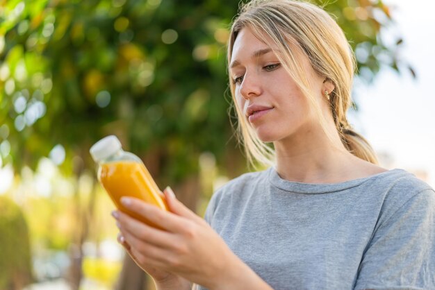 Junge blonde Frau hält draußen einen Orangensaft in der Hand