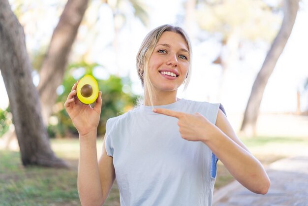 Foto junge blonde frau hält eine avocado im freien und zeigt darauf