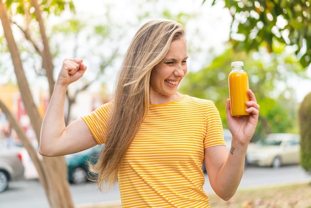 Junge blonde Frau hält einen Orangensaft im Freien und feiert einen Sieg