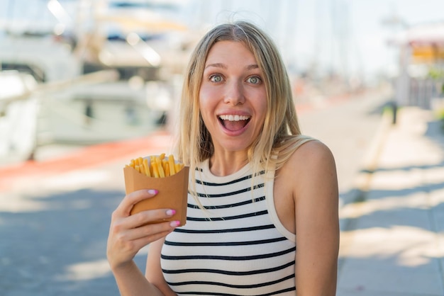 Junge blonde Frau hält mit Überraschung und schockiertem Gesichtsausdruck frittierte Chips im Freien in der Hand