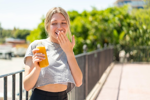 Junge blonde Frau hält mit überraschtem und schockiertem Gesichtsausdruck im Freien einen Orangensaft in der Hand