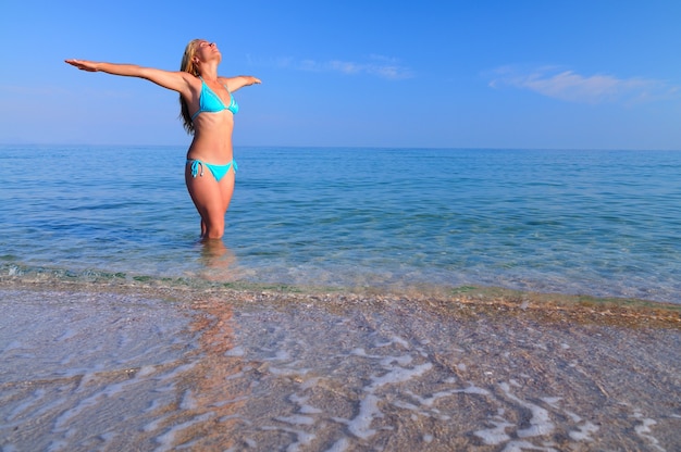 Junge blonde Frau im blauen Bikini, der im stillen Meerwasser steht und Sonnenschein am sonnigen Sommertag genießt. Konzept für Glück, Urlaub und Freiheit