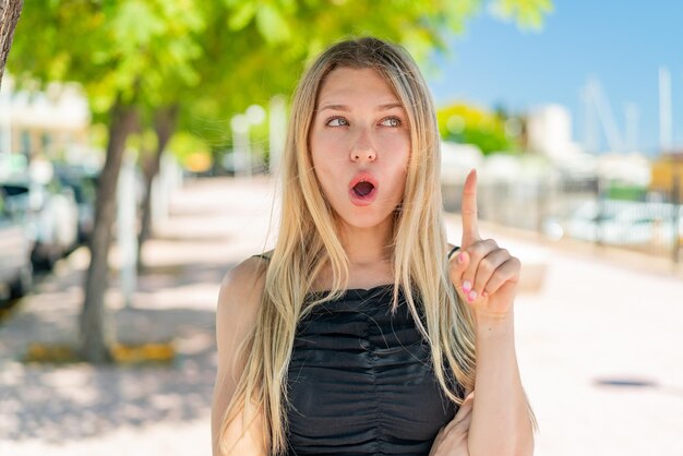 Junge blonde Frau im Freien denkt über eine Idee nach und zeigt mit dem Finger nach oben