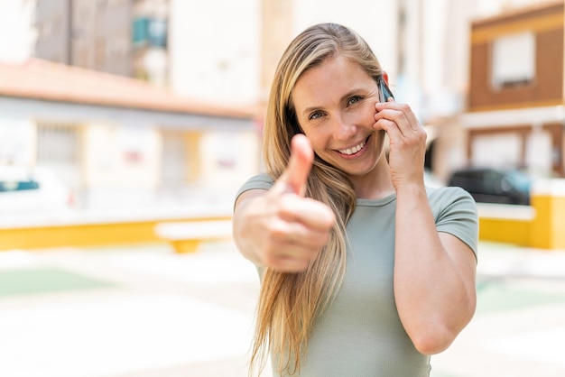 Foto junge blonde frau im freien, die ein gespräch mit dem handy führt und dabei den daumen nach oben zeigt