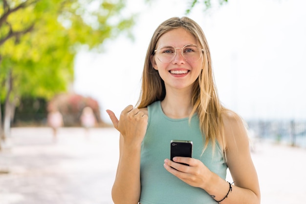 Foto junge blonde frau im freien, die ihr mobiltelefon benutzt und auf die seite zeigt