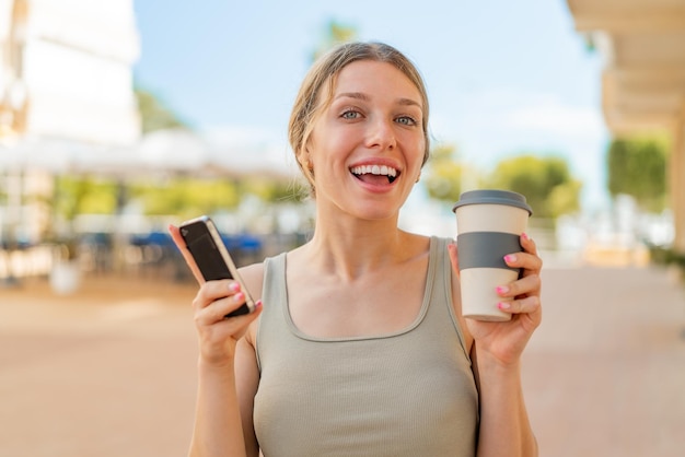 Junge blonde Frau im Freien, die ihr Mobiltelefon benutzt und mit glücklichem Gesichtsausdruck einen Kaffee in der Hand hält