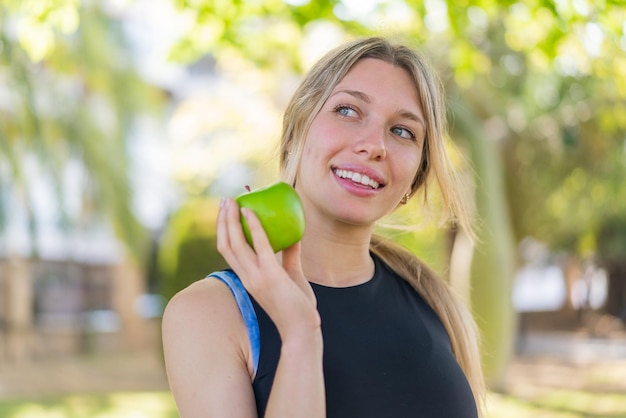 Junge blonde Frau im Freien hält einen Apfel mit glücklichem Gesichtsausdruck