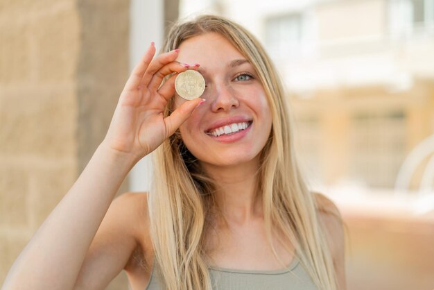 Junge blonde Frau im Freien hält einen Bitcoin mit glücklichem Gesichtsausdruck in der Hand