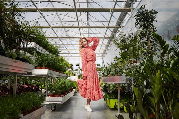 Junge blonde Frau im roten Kleid tanzt in ein Gewächshaus. Industrielles Gewächshaus mit Blumen in Töpfen rundum.
