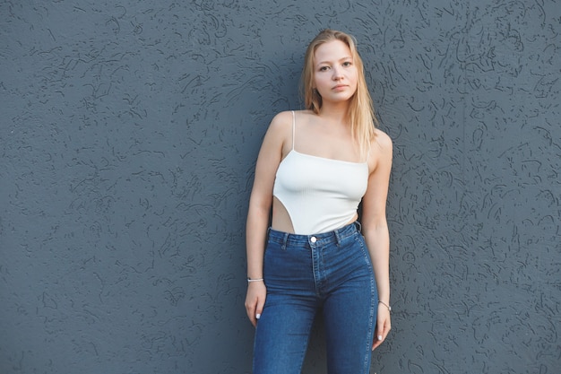 Junge blonde Frau im weißen ärmellosen T-Shirt und in den blauen Jeans, die über grauem Wandhintergrund stehen.