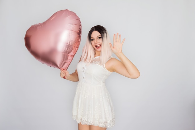 Junge blonde Frau im weißen Spitzenkleid, das großen rosa Herzballon in ihren Händen über weißem Hintergrund hält