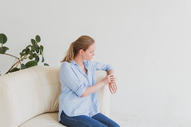 Junge blonde Frau in einem blauen Hemd sitzt auf der Couch in einem hellen Raum und schaut auf seine Uhr