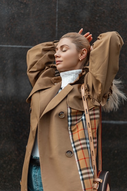 junge blonde Frau in einem Pullover und einem beigen Trenchcoat, der nahe einem schwarzen modernen Gebäude aufwirft