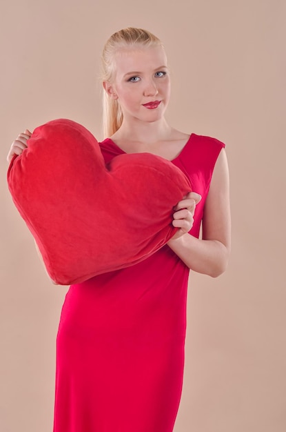 Junge blonde Frau in einem roten Kleid hält ein rotes Herz in ihren Händen auf einem beigen Hintergrund