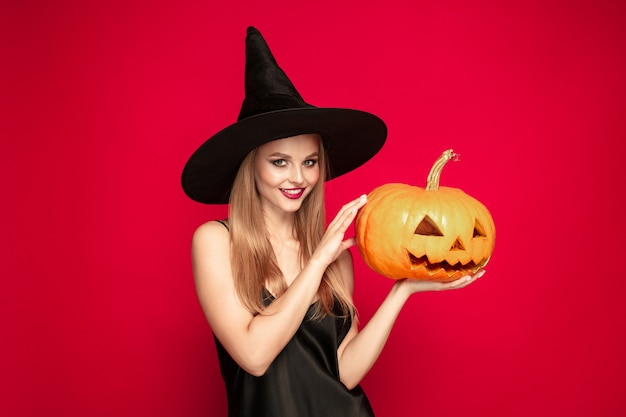 Junge blonde Frau in schwarzem Hut und Kostüm auf rotem Grund. Attraktives kaukasisches weibliches Modell. Halloween, schwarzer Freitag, Cyber-Montag, Verkauf, Herbstkonzept. Exemplar. Hält Kürbis, lächelt.