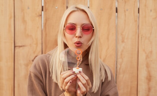 Junge blonde Frau in warmer Kleidung und trendiger rosa Sonnenbrille, die Spaß hat und Seifenblase bläst, während sie gegen hölzernen Plankenzaun steht