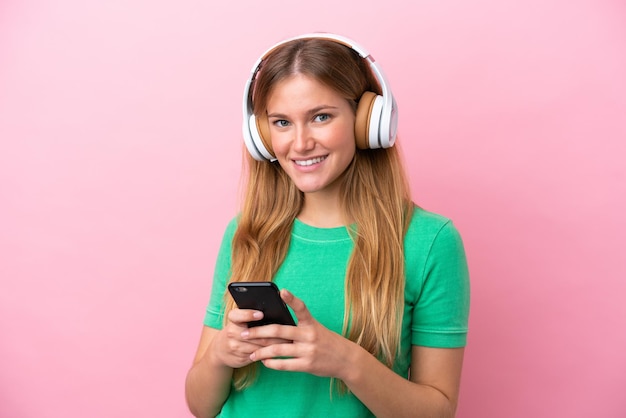 Junge blonde Frau isoliert auf rosafarbenem Hintergrund, die Musik mit einem Handy hört und nach vorne schaut