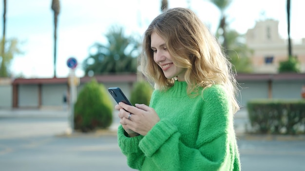 Junge blonde Frau lächelt selbstbewusst mit Smartphone auf der Straße
