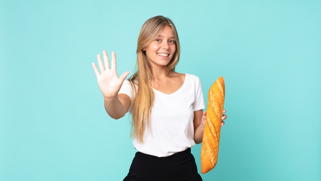 Junge blonde Frau lächelt und sieht freundlich aus, zeigt Nummer fünf und hält ein Brotbaguette