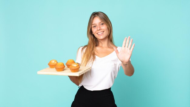 Junge blonde Frau lächelt und sieht freundlich aus, zeigt Nummer fünf und hält einen Muffins-Troy