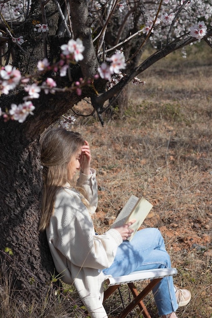 Junge blonde Frau liest ein Buch, das auf dem Feld unter einem Baum mit Blumen sitzt