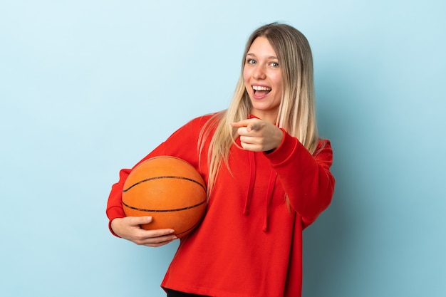 Junge blonde Frau lokalisiert auf blauer Wand, die Basketball spielt und nach vorne zeigt