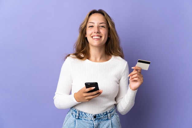 Junge blonde Frau lokalisiert auf lila Wand, die mit dem Handy mit einer Kreditkarte kauft