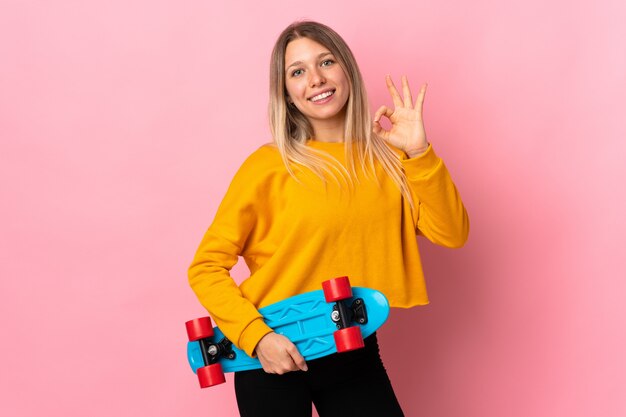 Junge blonde Frau lokalisiert auf rosa Wand mit einem Schlittschuh und tut OK Zeichen