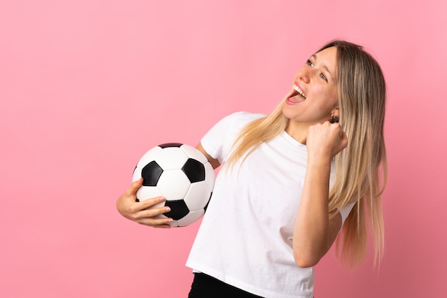 Junge blonde Frau lokalisiert auf rosa Wand mit Fußball, der einen Sieg feiert