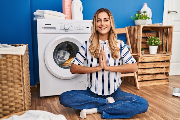 Junge blonde Frau macht Yoga-Übungen und wartet in der Waschküche auf die Waschmaschine
