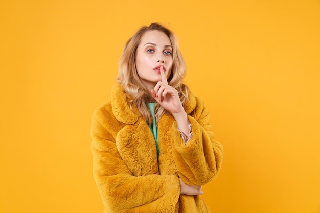 Junge blonde Frau Mädchen im gelben Pelzmantel posiert isoliert auf orangefarbenem Hintergrund Studioporträt. People-Lifestyle-Konzept. Mock-up-Kopienbereich. Schweigen zu sagen, sei ruhig mit dem Finger auf den Lippen, shhh Geste.