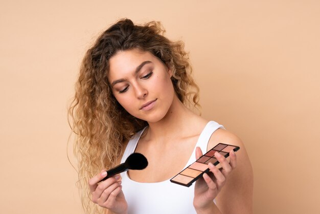 Junge blonde Frau mit dem gelockten Haar lokalisiert auf Beige mit Make-up-Palette