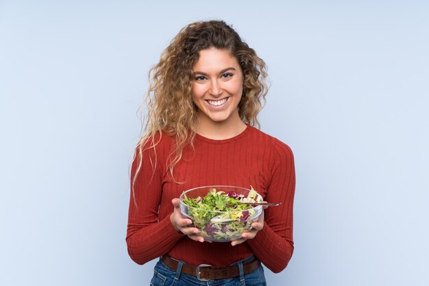 Junge blonde Frau mit dem lockigen Haar, das einen Salat über Wand hält