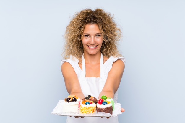 Junge blonde Frau mit dem lockigen Haar, das Lots verschiedene Minikuchen anhält