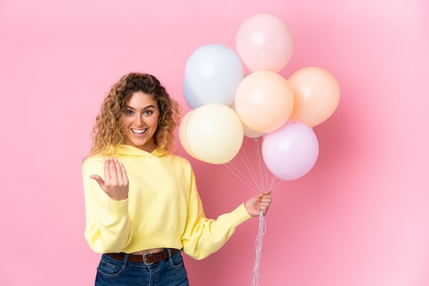 Junge blonde Frau mit dem lockigen Haar, das viele Luftballons auf Rosa einlädt, einladend zu kommen